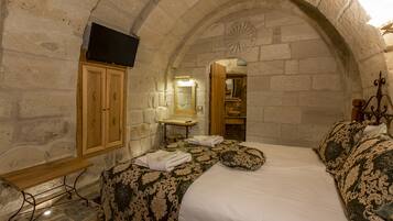 A stone-walled bedroom with a bed, wooden cabinet, and a flat-screen TV mounted on the wall.
