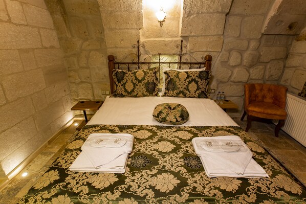Una cama bien hecha con una colcha verde y dorada, dos toallas blancas y una almohada decorativa en una habitación con pared de piedra.
