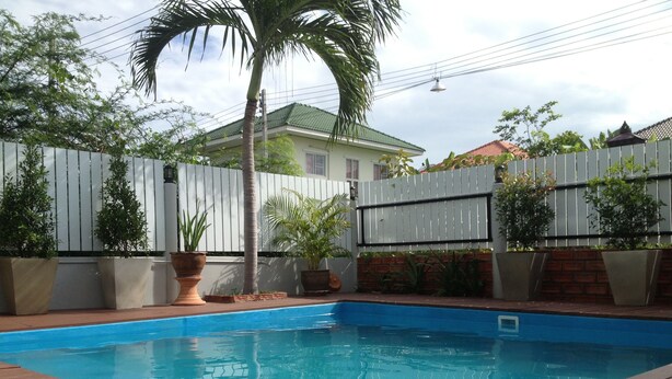 Una piscina con cubierta de madera, rodeada por una valla blanca, macetas con plantas y una palmera.