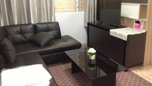 Un salón con un sofá de cuero negro, una mesa de centro de vidrio, un televisor sobre un mueble negro y una pequeña mesa blanca con un juguete rosa.