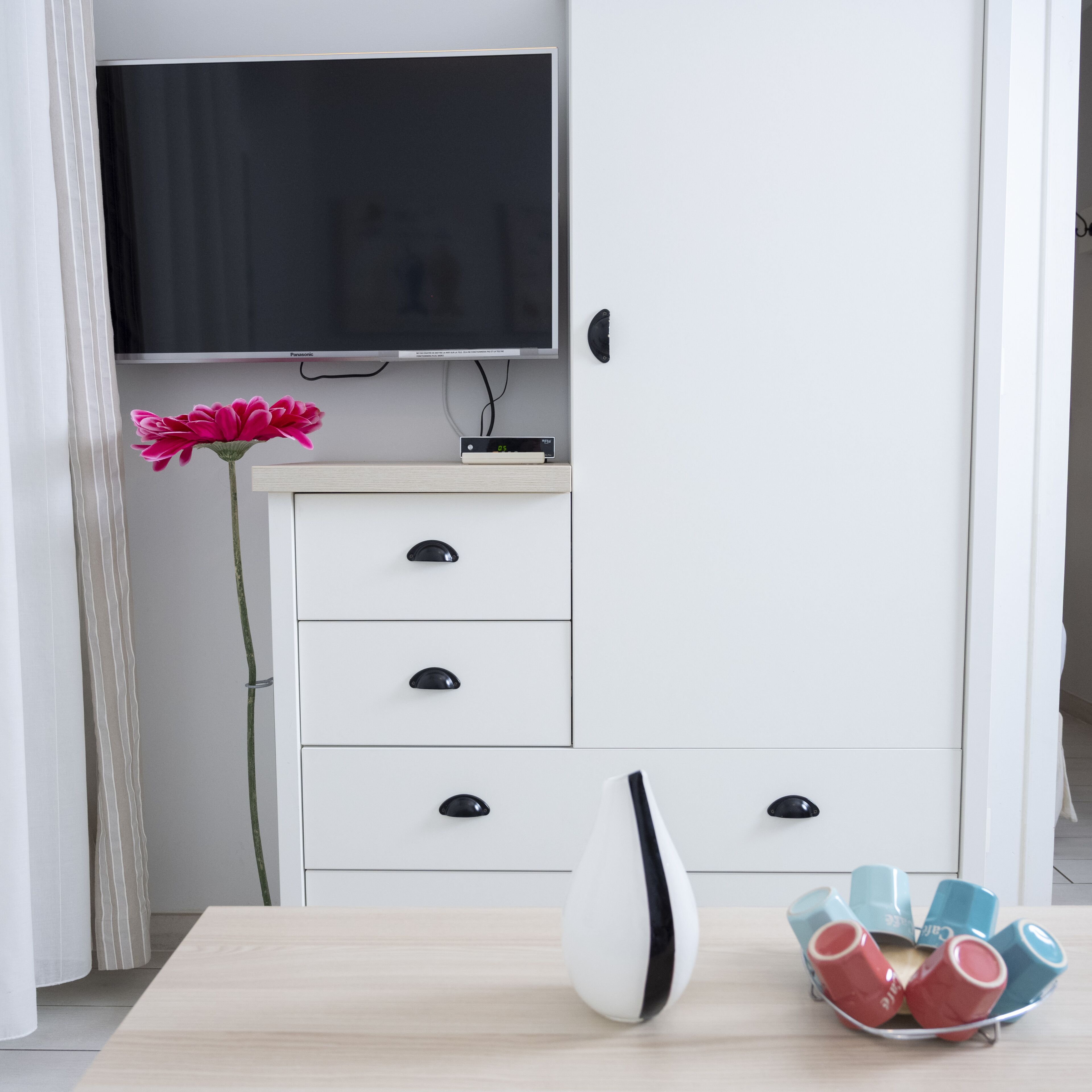 Una habitación con una televisión de pantalla plana montada en la pared, un mueble blanco con cajones, una mesa con un jarrón blanco y un cuenco con tazas de colores, y una flor rosa a la izquierda.