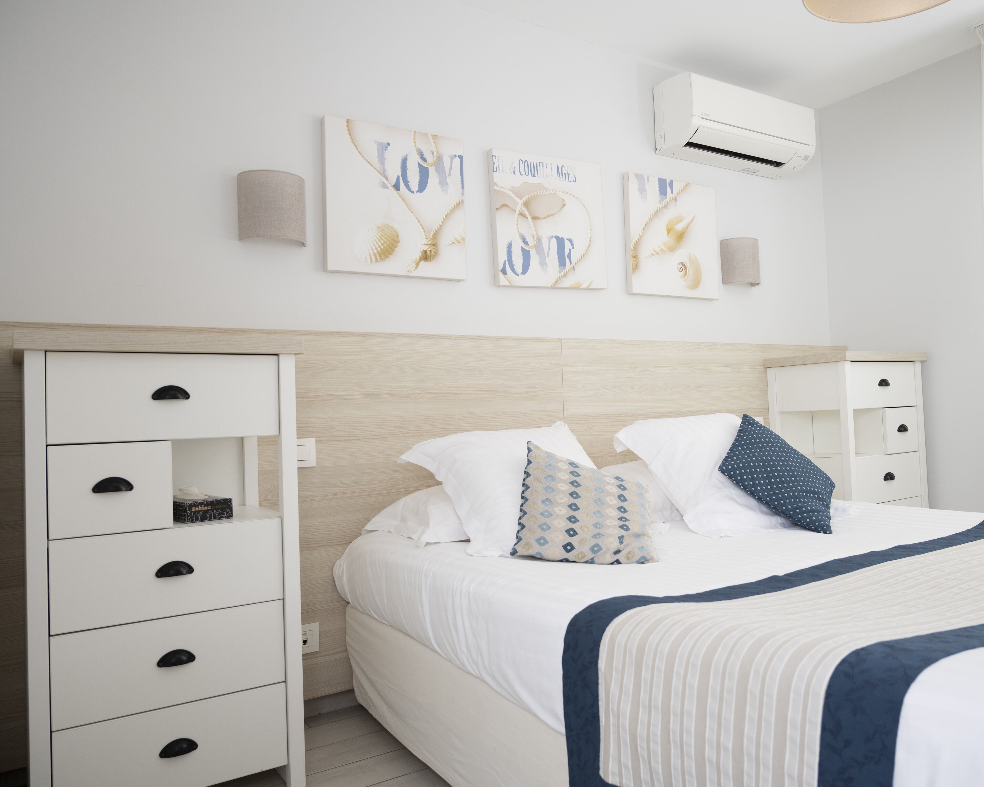 A modern bedroom with a wooden headboard, a white bed with multiple pillows, a bedside table, and wall-mounted artwork.
