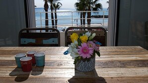 A wooden table with a vase of flowers and four cups labeled "Café" in front of a window overlooking a beach with palm trees.