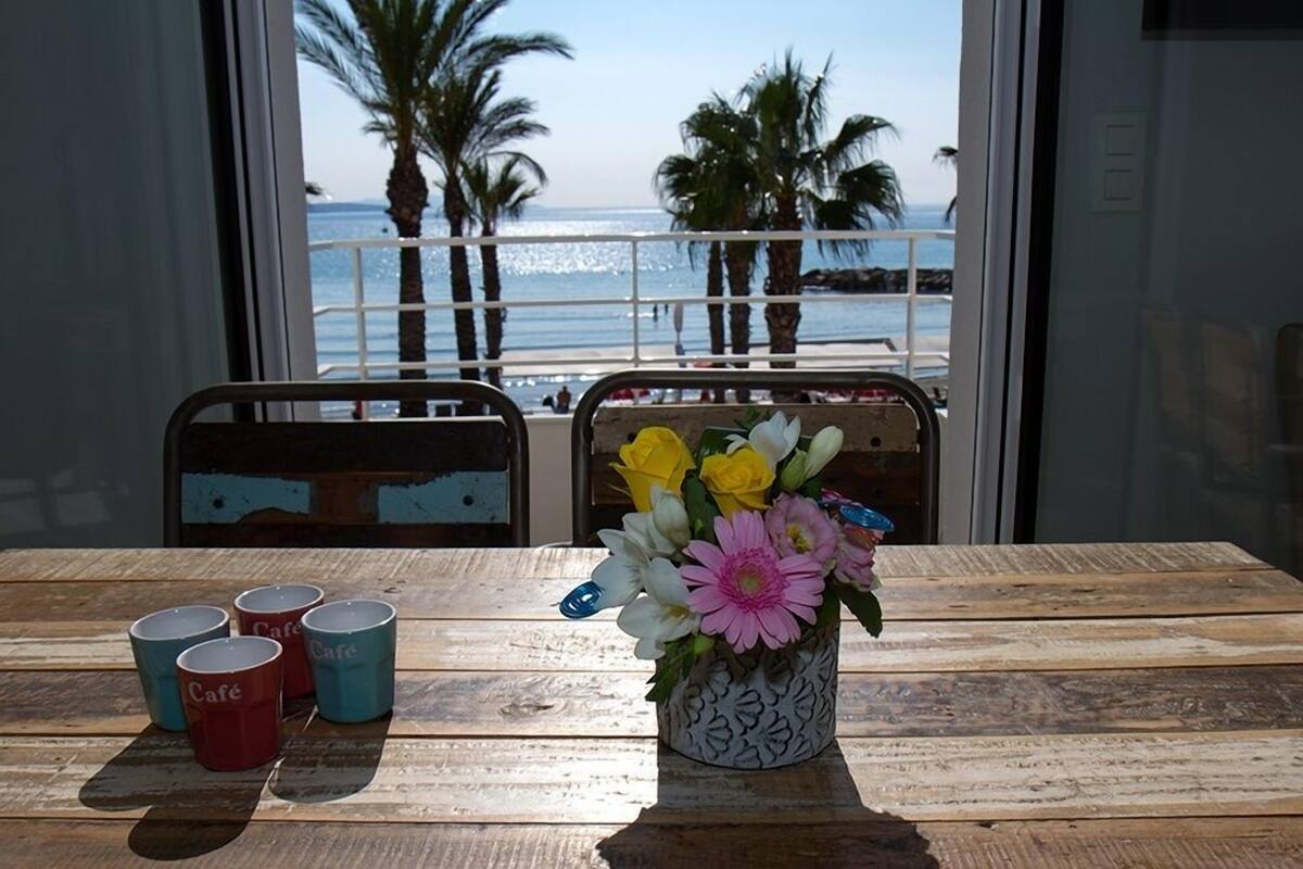 Taula de fusta amb un ram de flors i quatre tasses amb la paraula "Café" davant d'una finestra que donen vistes a una platja amb palmeres.