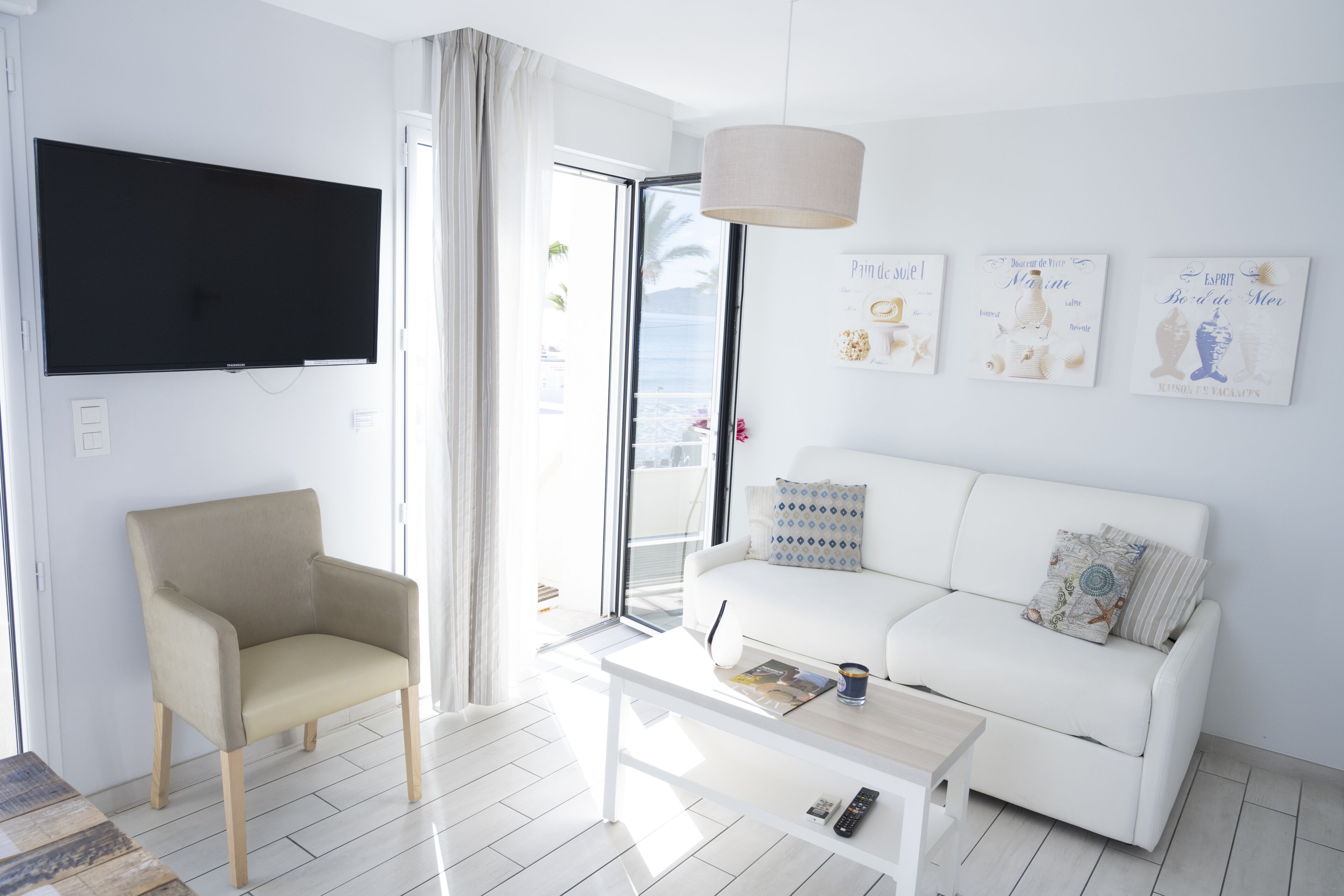 Ein modernes Wohnzimmer mit einem weißen Sofa, einem beigen Sessel und einem Couchtisch. An der Wand ist ein Flachbildfernseher angebracht, und ein Fenster mit Vorhängen vorhanden.