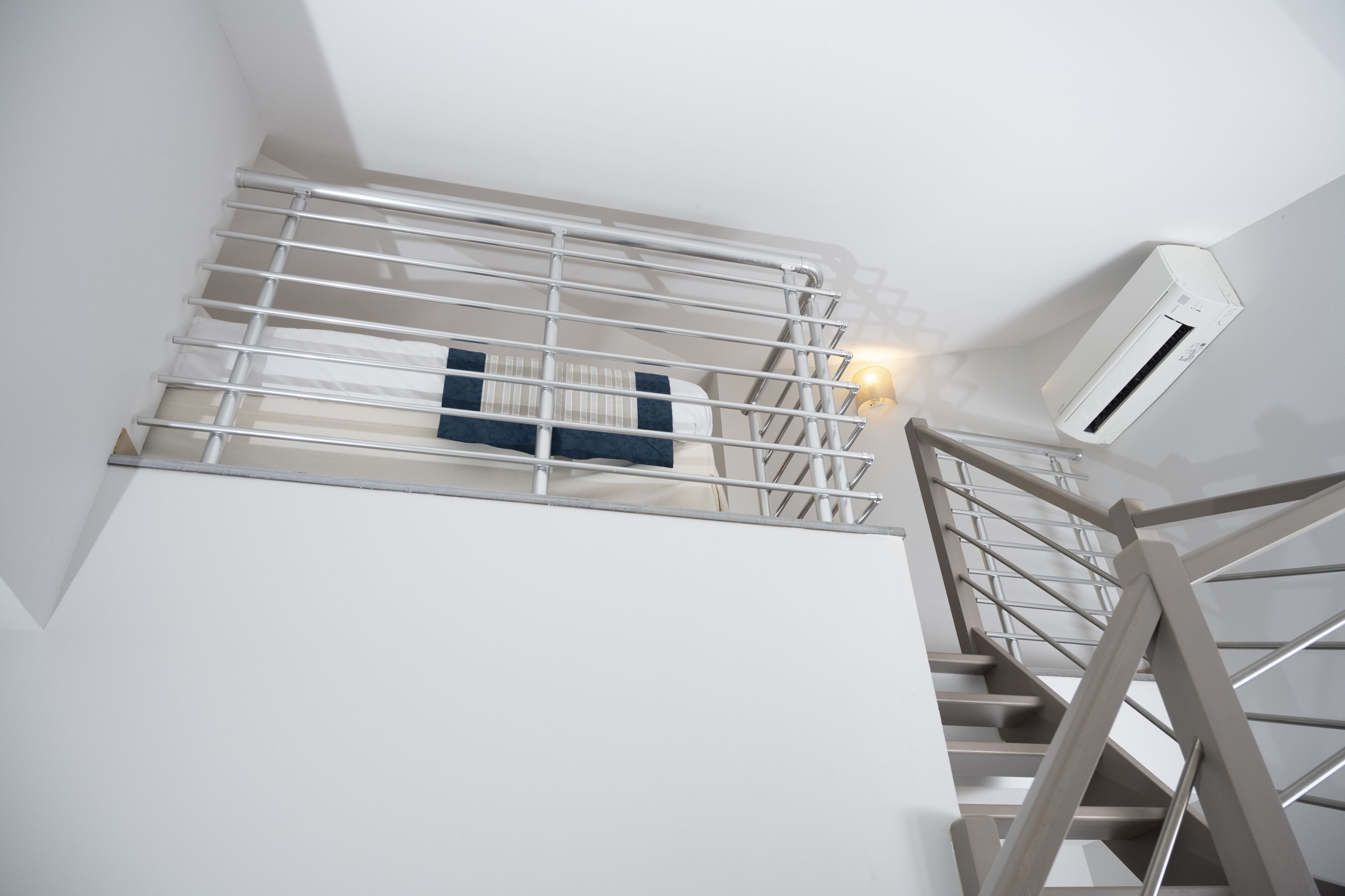 A modern staircase with metal railings and a wooden handrail, leading to a room with a bed and an air conditioning unit mounted on the wall.
