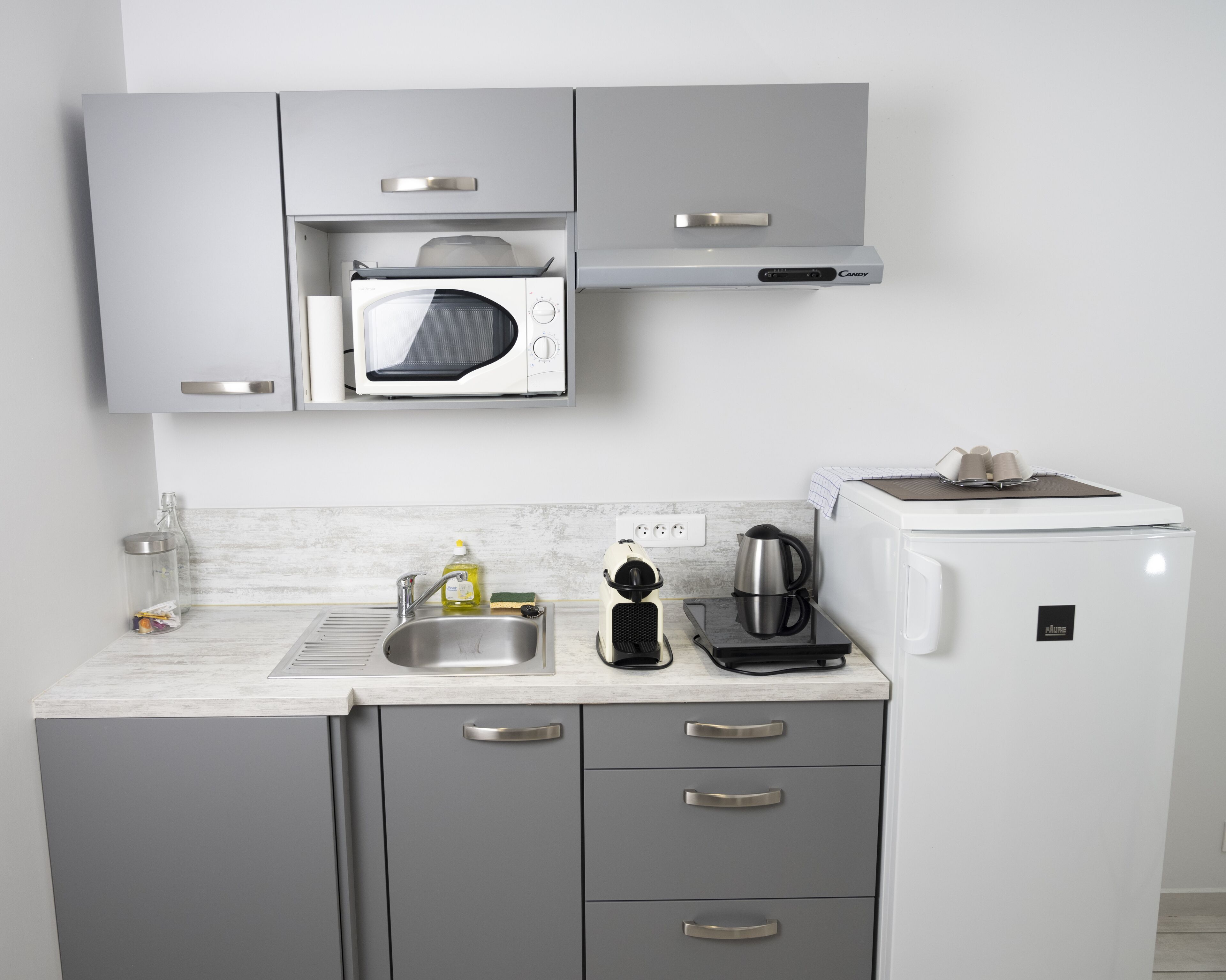 Una cocina compacta con gabinetes grises, un microondas, una cafetera y un refrigerador pequeño.