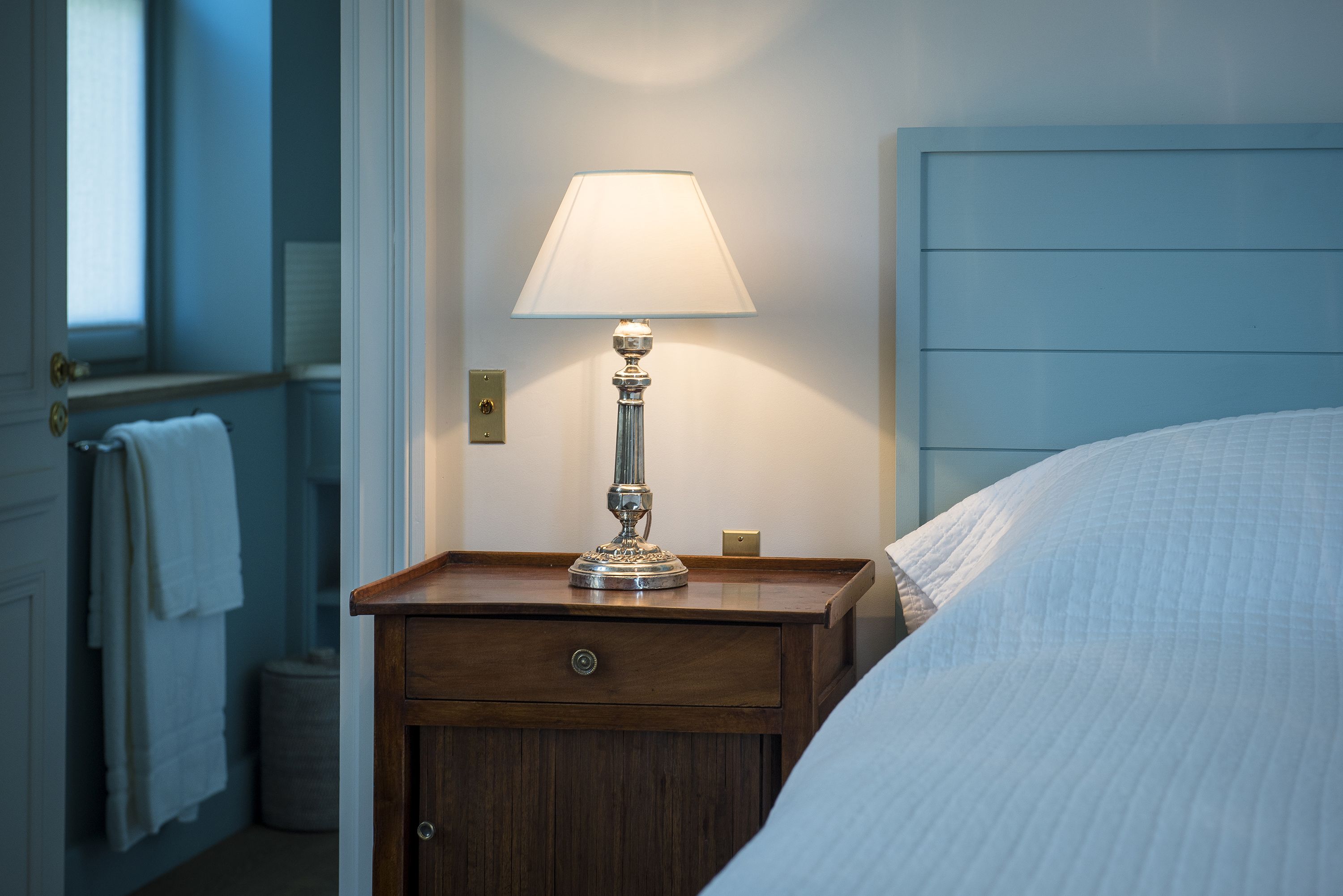 A wooden bedside table with a lamp, a bed with white bedding, and a blue headboard.