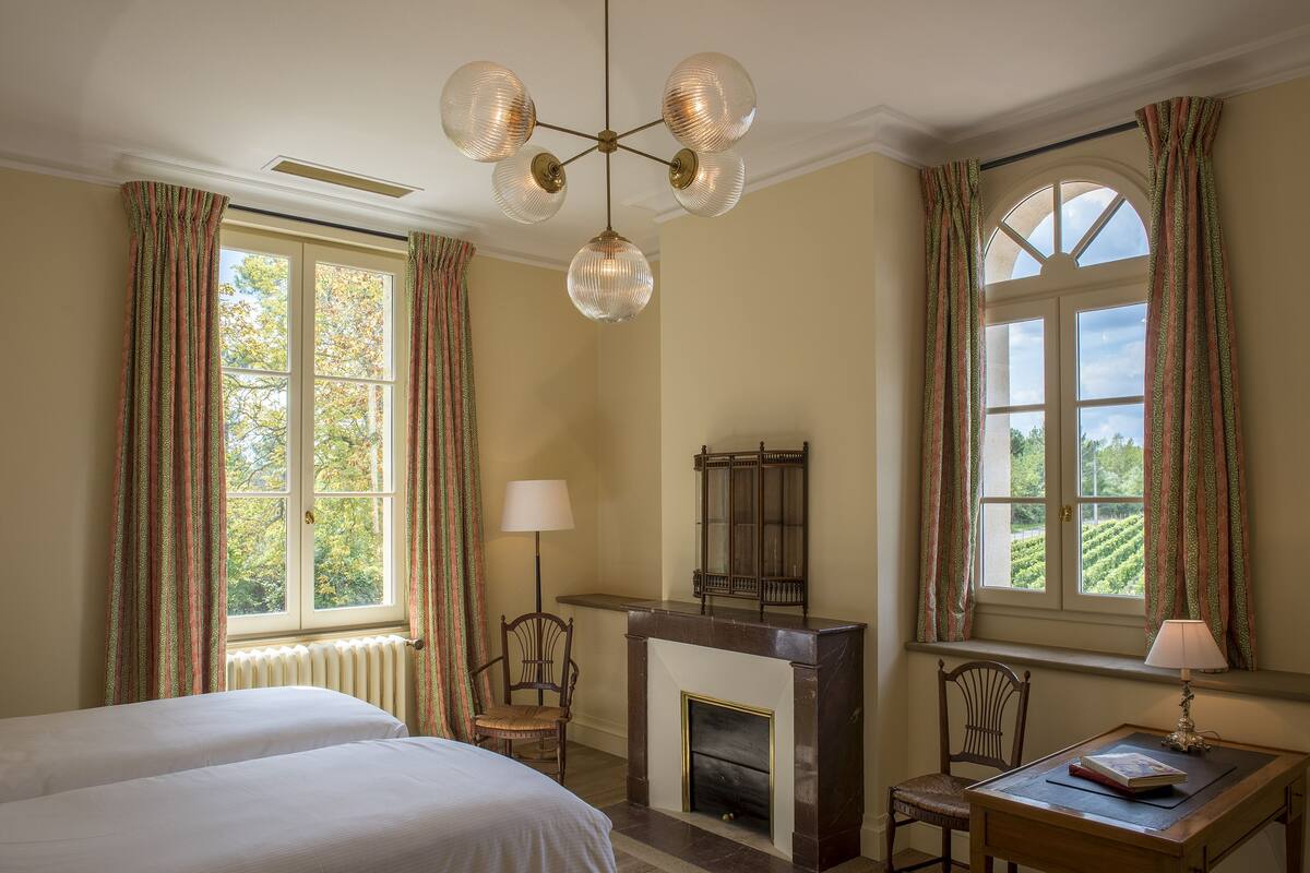 A room with two beds, a fireplace, a desk with a lamp, and a view of the outdoors.