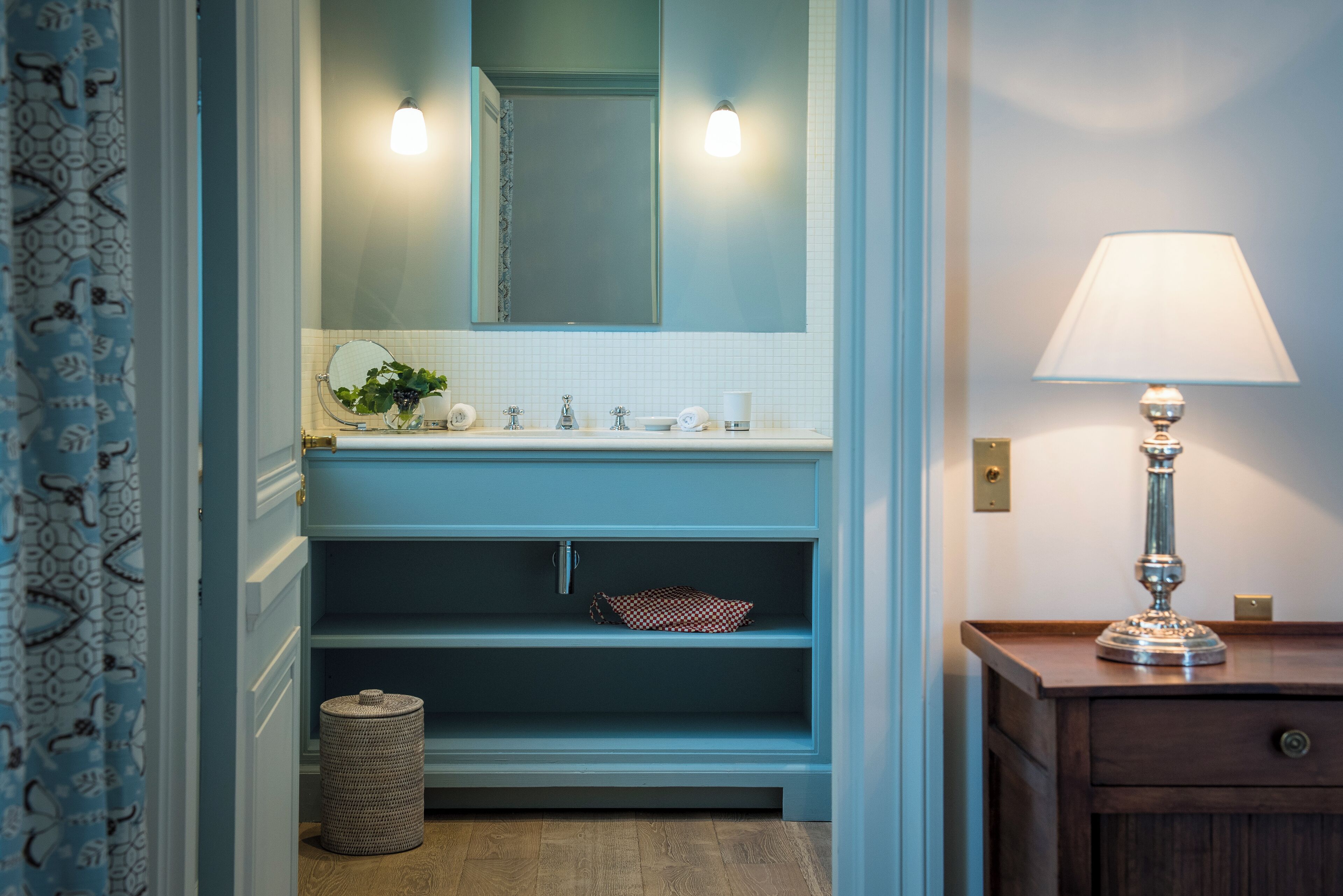 A bathroom with a blue vanity, a mirror, a sink, a towel rack, a small trash bin, and a wooden nightstand with a lamp.