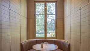 A corner seating area with a round table, a bottle of wine, and glasses, surrounded by wooden panel walls and a large window with a view of greenery. - Haut-Bailly - La Chartreuse - Anciennement Château Le Pape (Leognan)