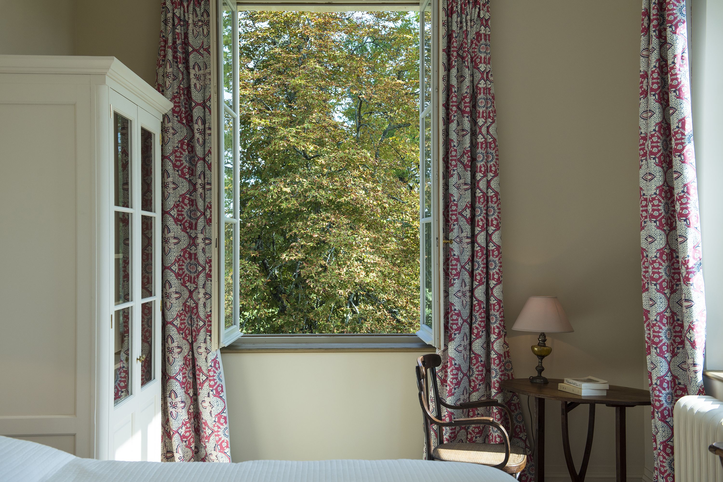 A bedroom with a large window, a chair, a bedside table with a lamp, and a radiator.