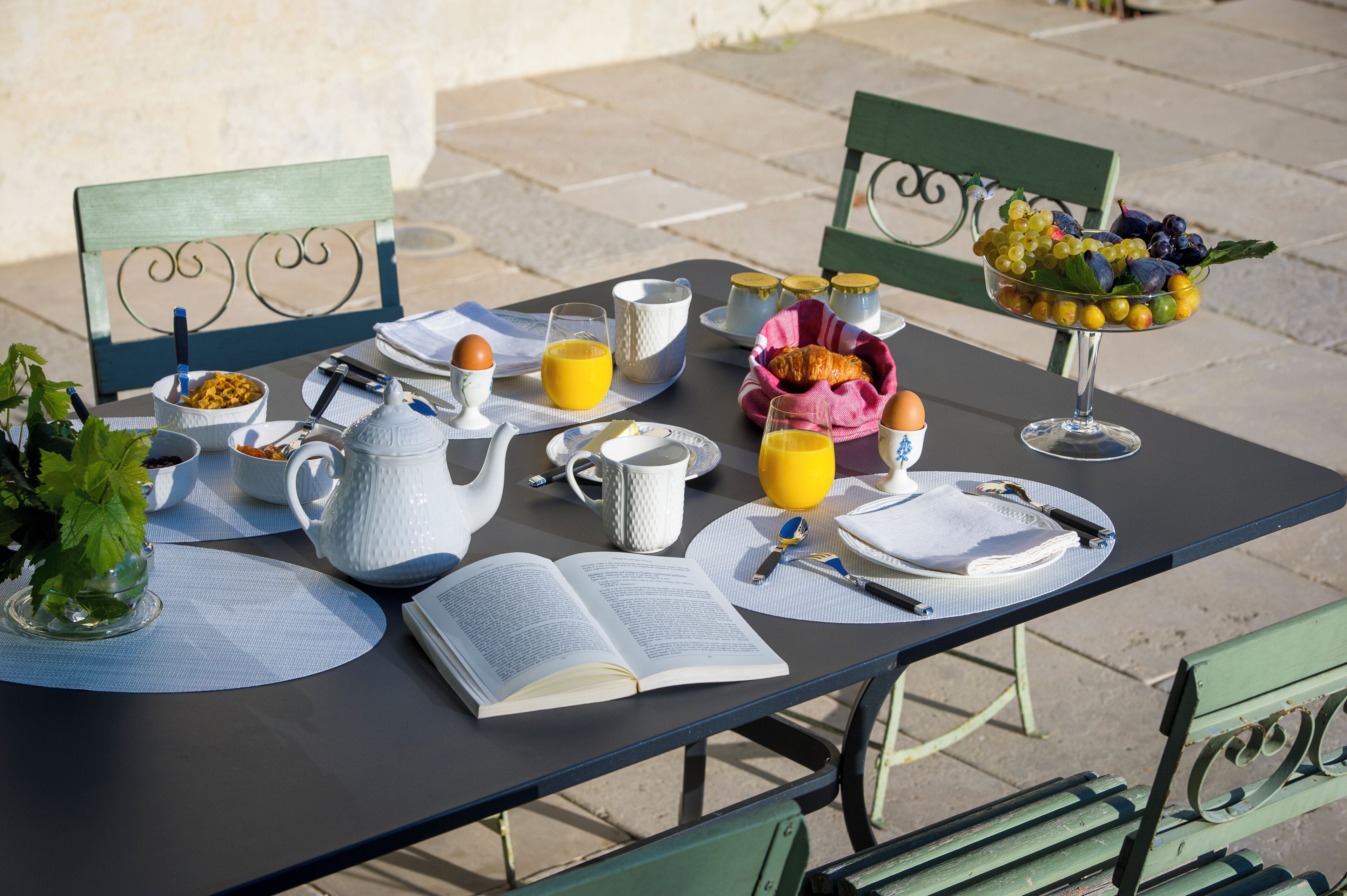 A breakfast table set with a book, a teapot, cups, a bowl of fruit, and a glass of orange juice.