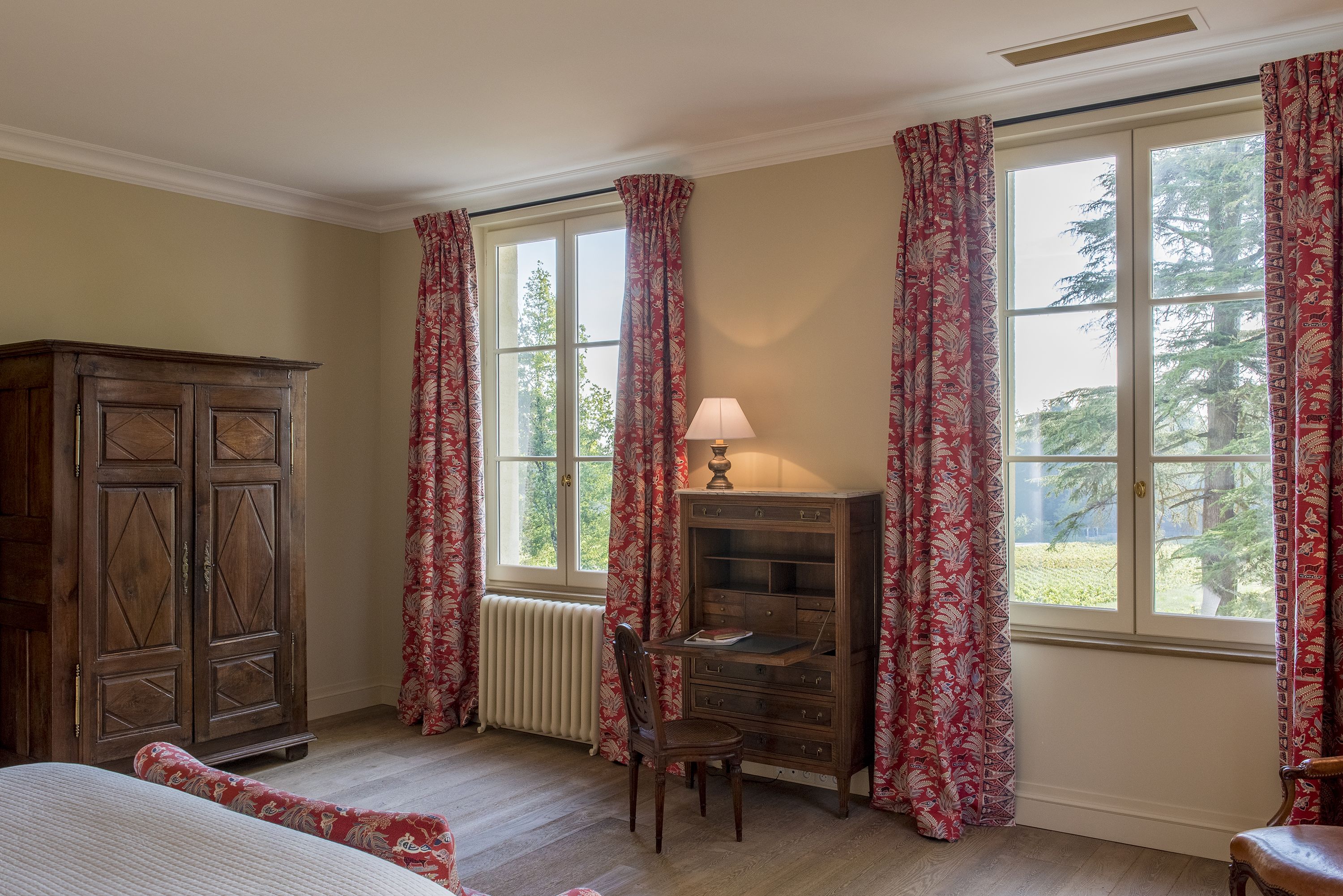 A room with a wooden cabinet, a desk with a lamp, and a chair. There are large windows with curtains.