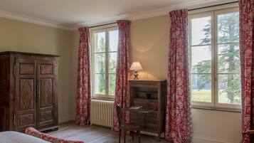 A room with a wooden cabinet, a desk with a lamp, and a chair. There are large windows with curtains.