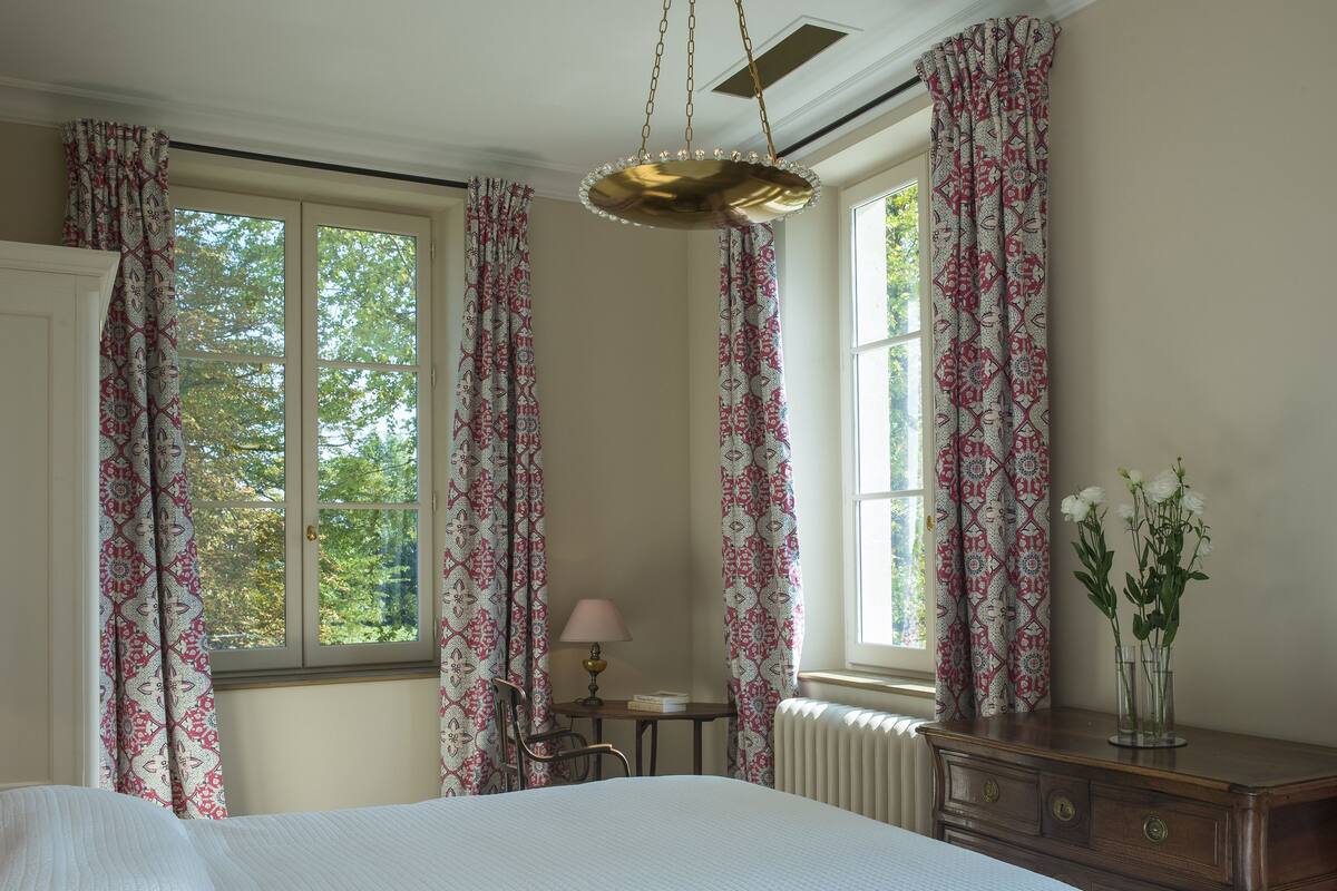 A bedroom with a bed, a desk with a lamp, a dresser, and a window with curtains.