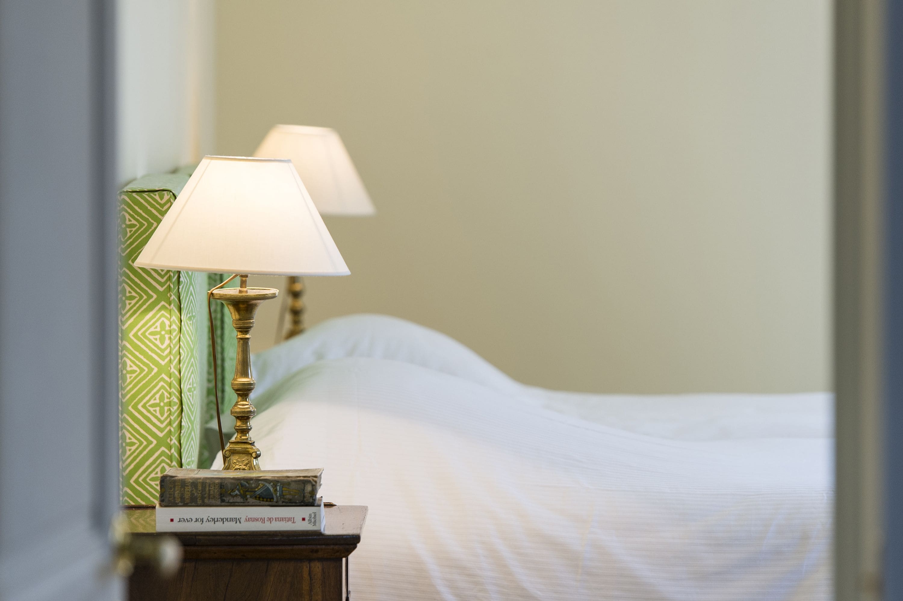 A neatly made bed with white linens, a bedside lamp with a green patterned lampshade, and a book on the nightstand.