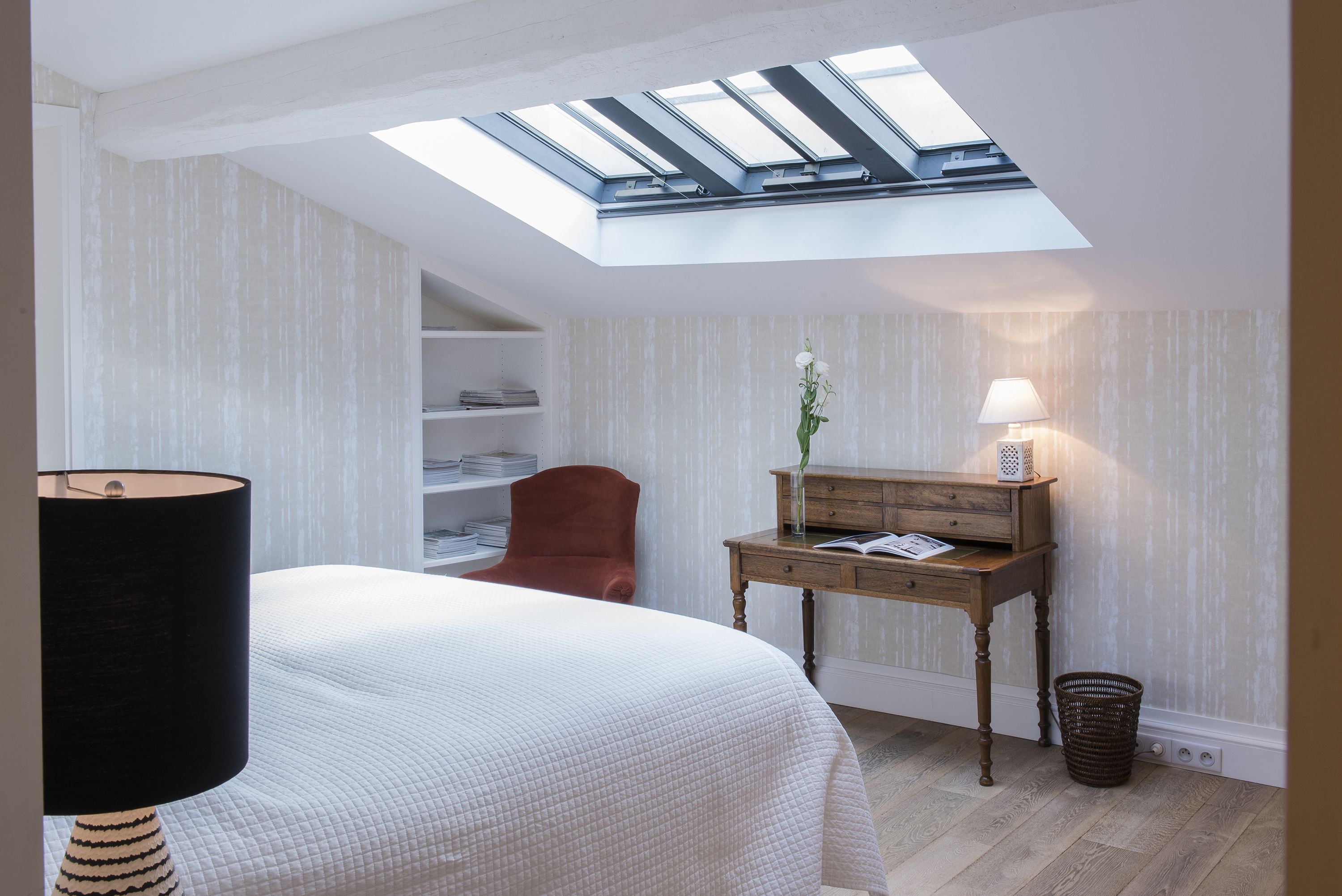 A bedroom with a skylight, a bed, a desk, a chair, and a lamp.