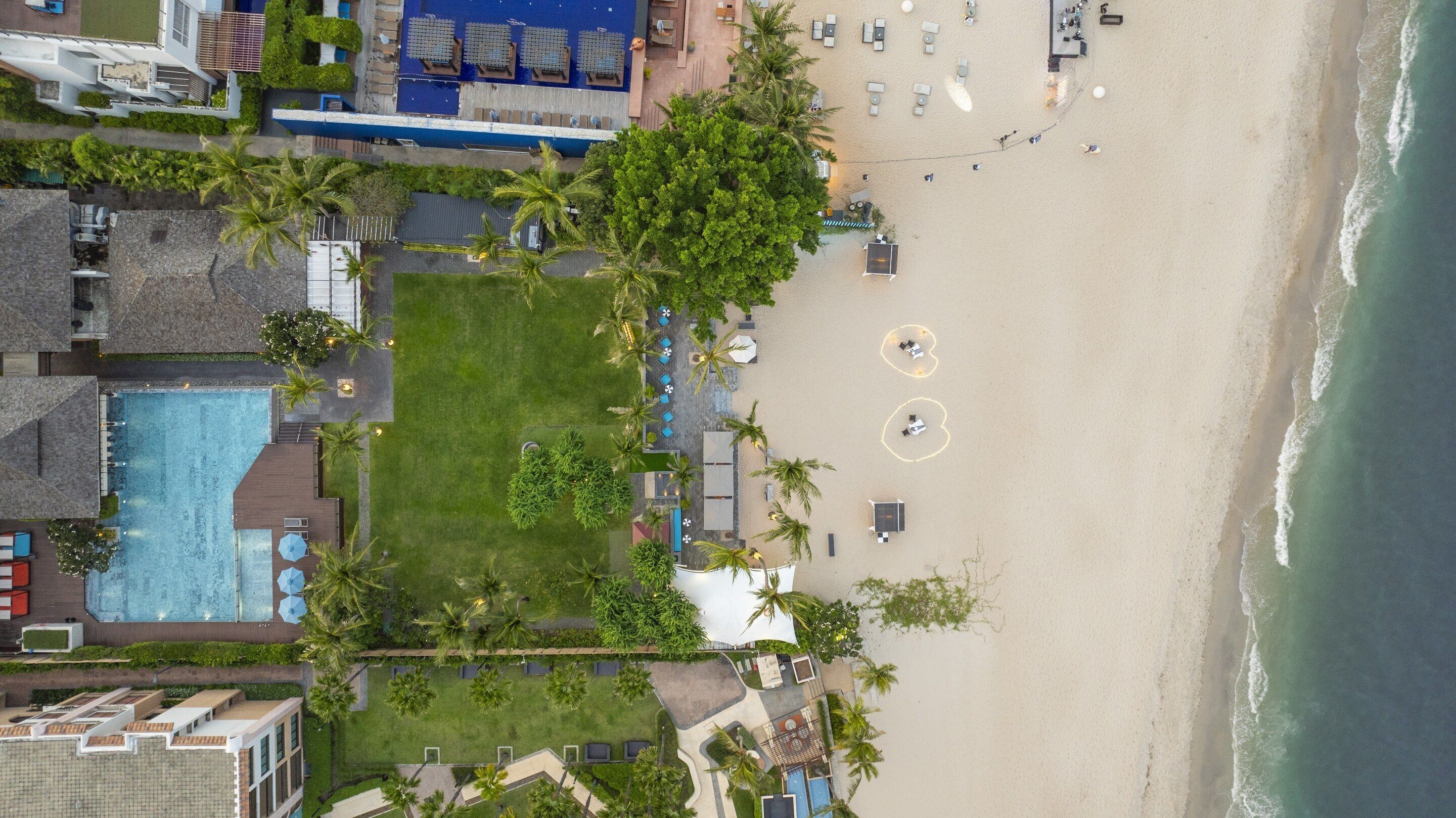 Vista aerea di un resort sulla spiaggia con piscina, lettini e palme.