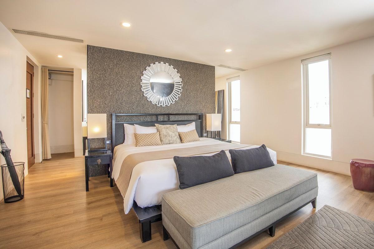 A modern hotel room with a large bed, a bench, a bedside table, and a mirror on the wall.