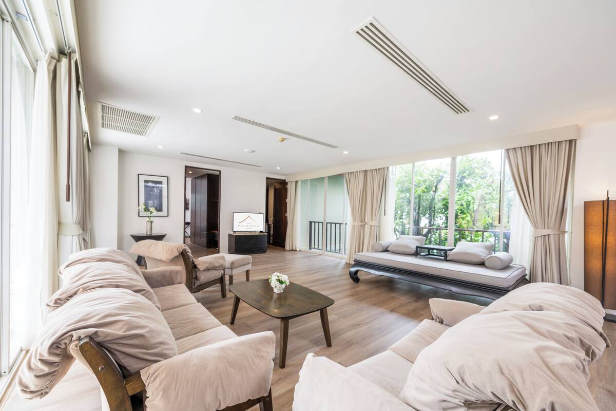 A spacious living room with a sofa set, a coffee table, and a TV. There is a balcony with a view of greenery.