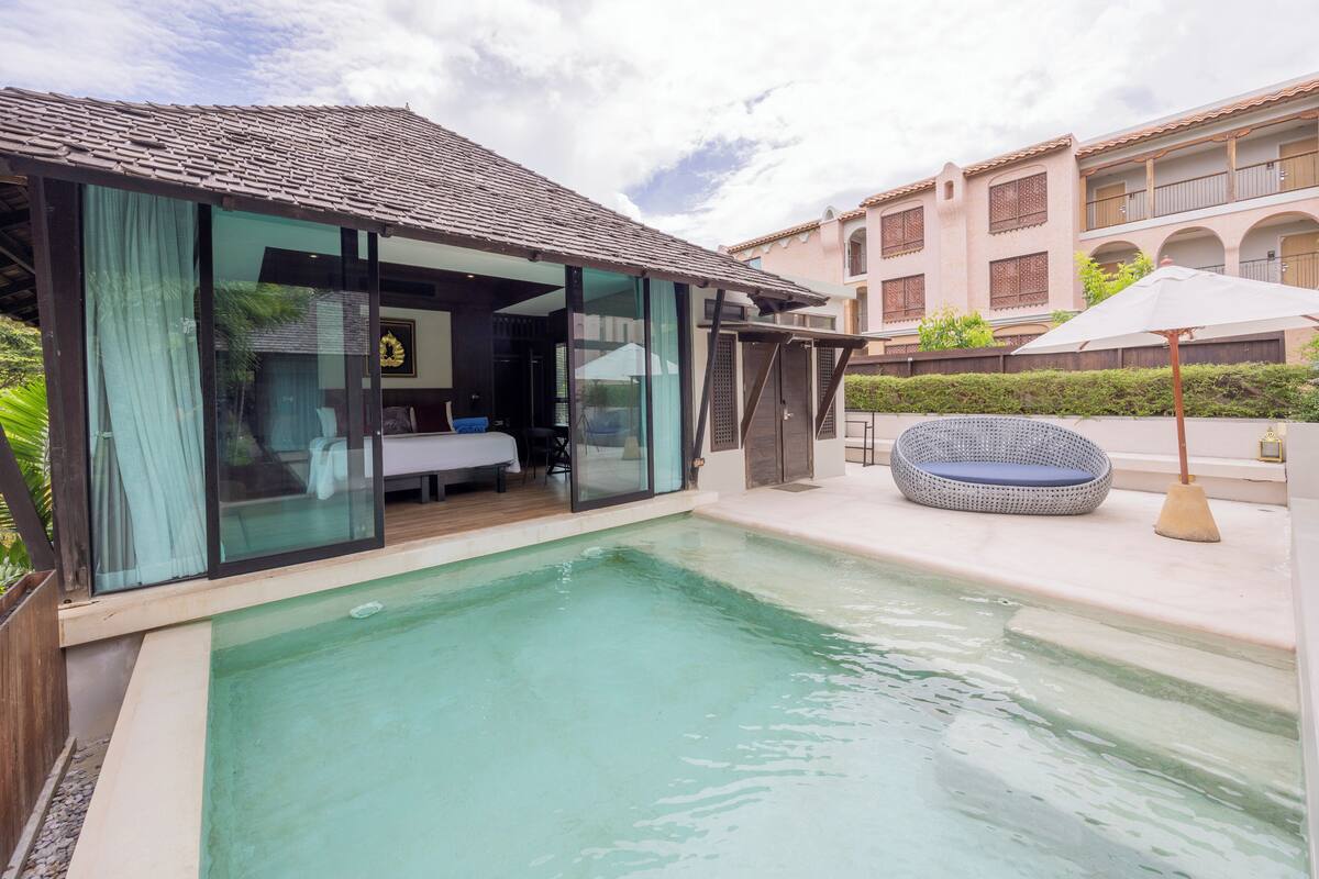 A modern pool area with a glass-enclosed room, outdoor seating, and a building with balconies.
