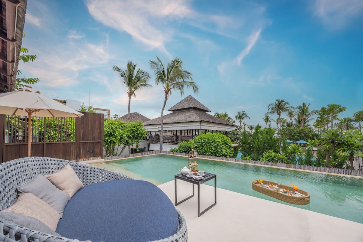 A poolside area with a wicker lounge chair, a small table with a tray of snacks, and a view of a resort building with palm trees.