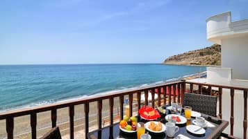 A breakfast table with plates, cups, and a fruit basket on a balcony overlooking the sea.
