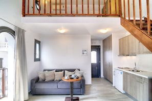 Een modern appartement met een grijze bank, een klein rond tafeltje, een keuken met houten kastjes en een trap.
