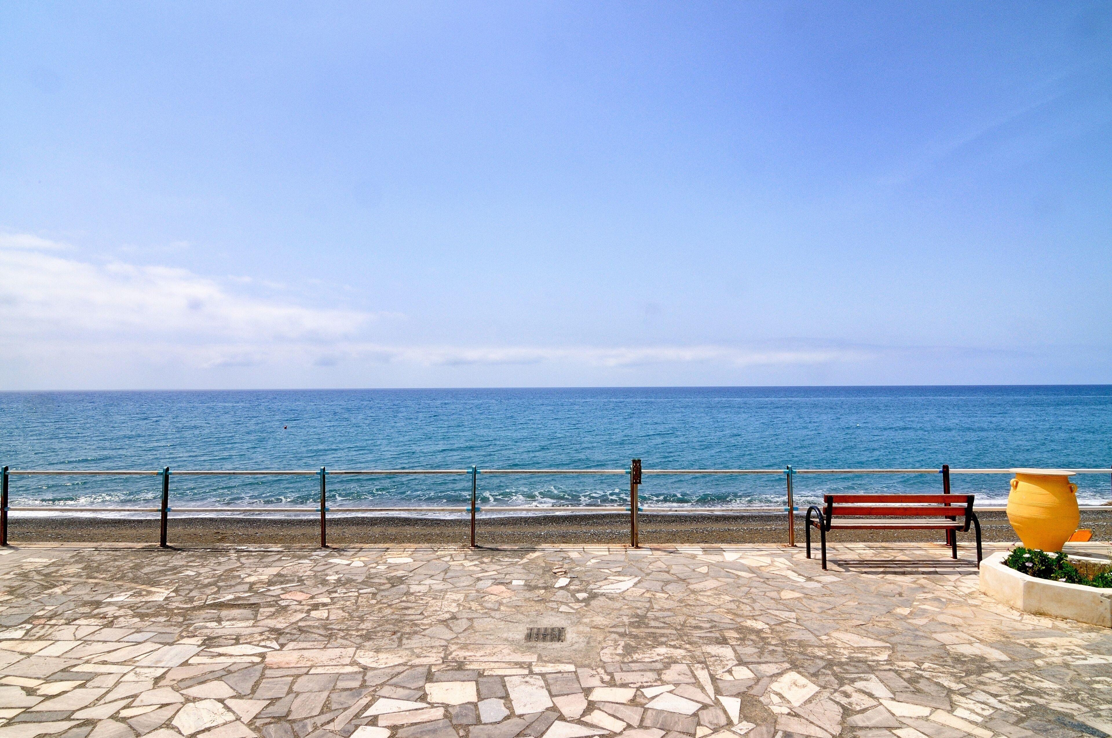 A stone-paved promenade with a bench and a yellow pot, overlooking a calm sea.