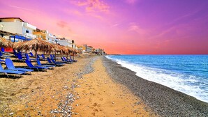 A beach with rows of blue lounge chairs and thatched umbrellas, a pebbly beach, and buildings in the background. - Myrtos Mare Suites (Ierapetra)