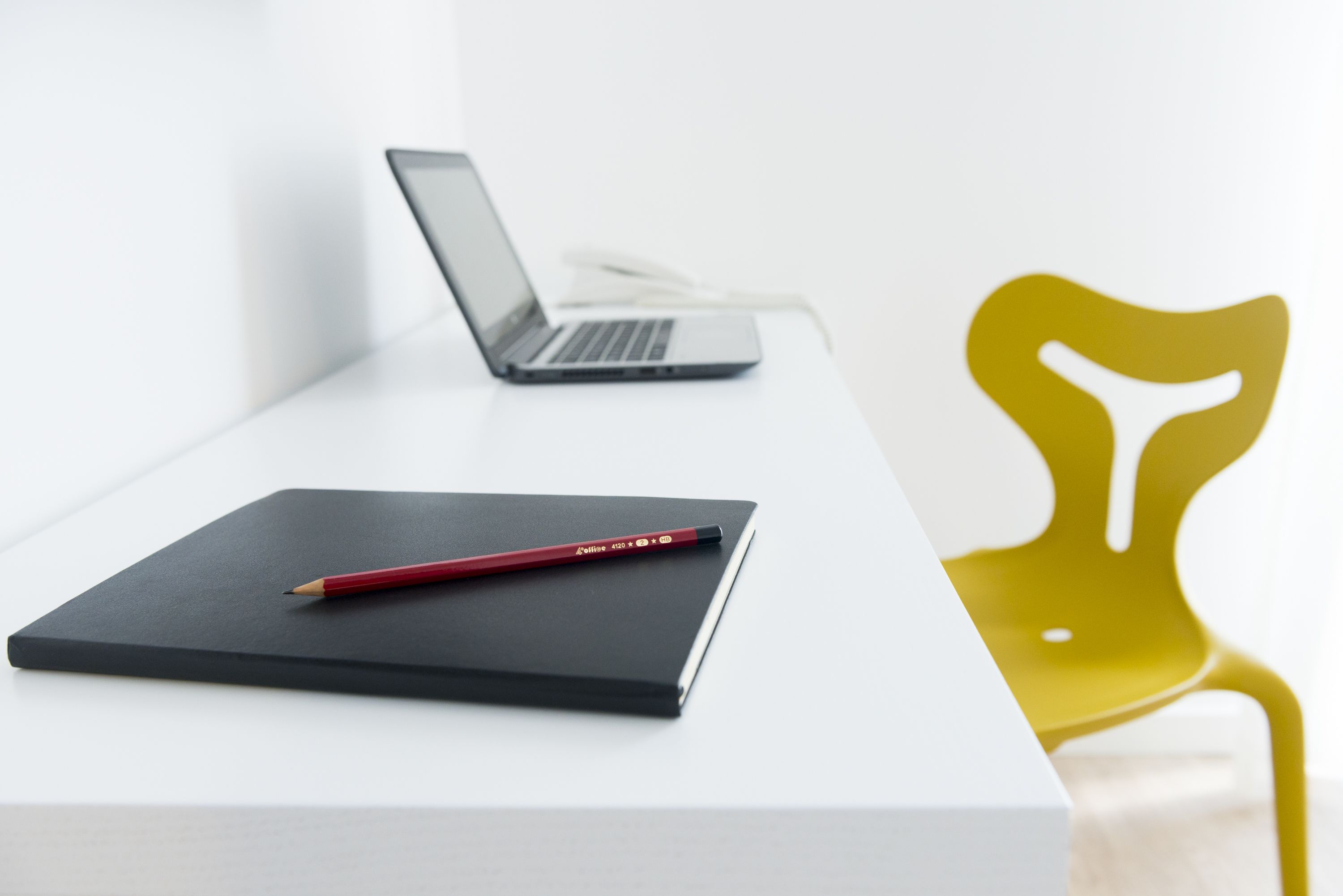 A white desk with a laptop, a red pencil, and a black notebook.