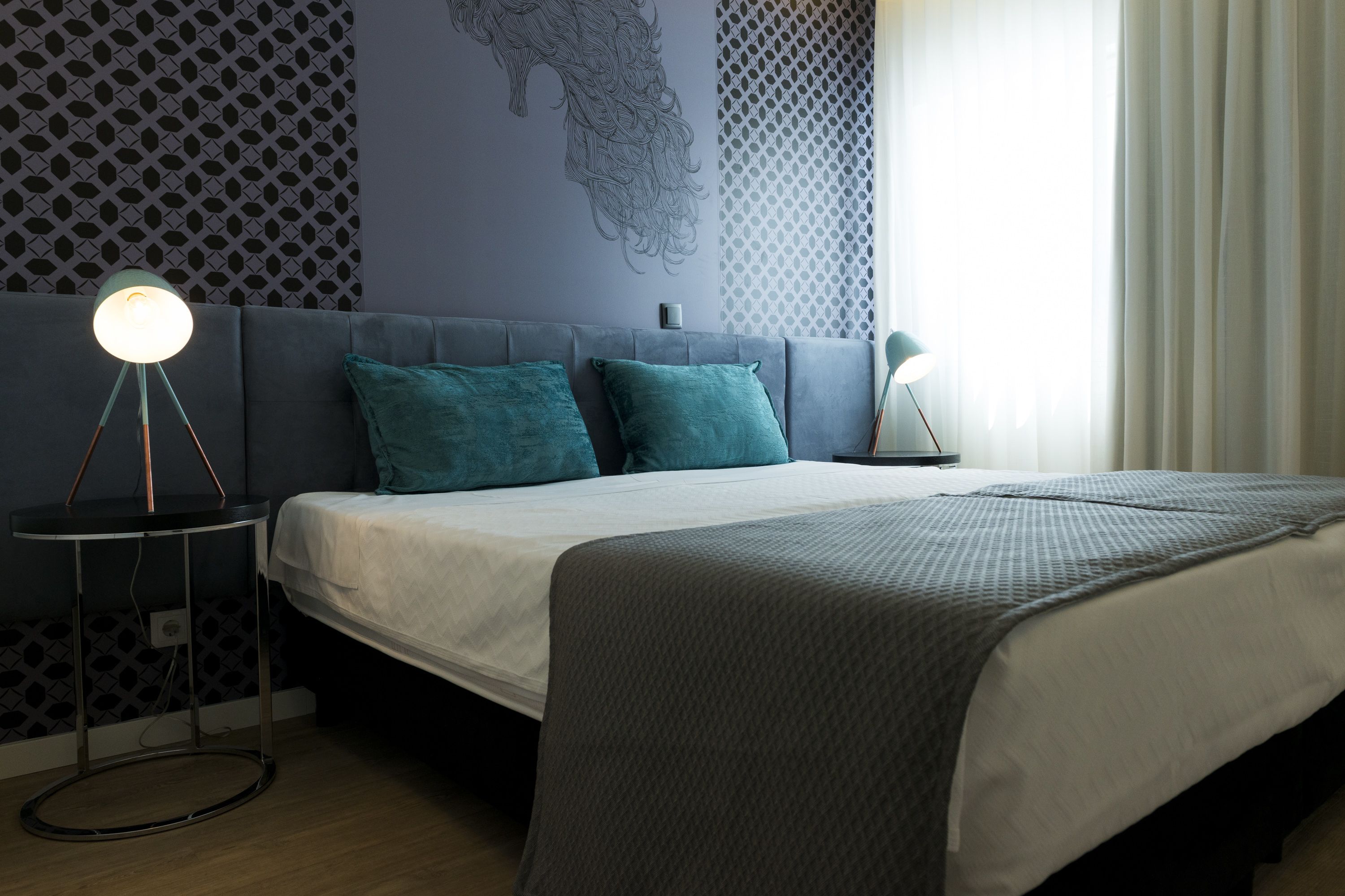 A modern bedroom with a grey sofa bed, teal pillows, a white duvet, and a patterned wall.