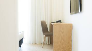 A hotel room with a wooden desk, a chair, a telephone, and a large window with white curtains.