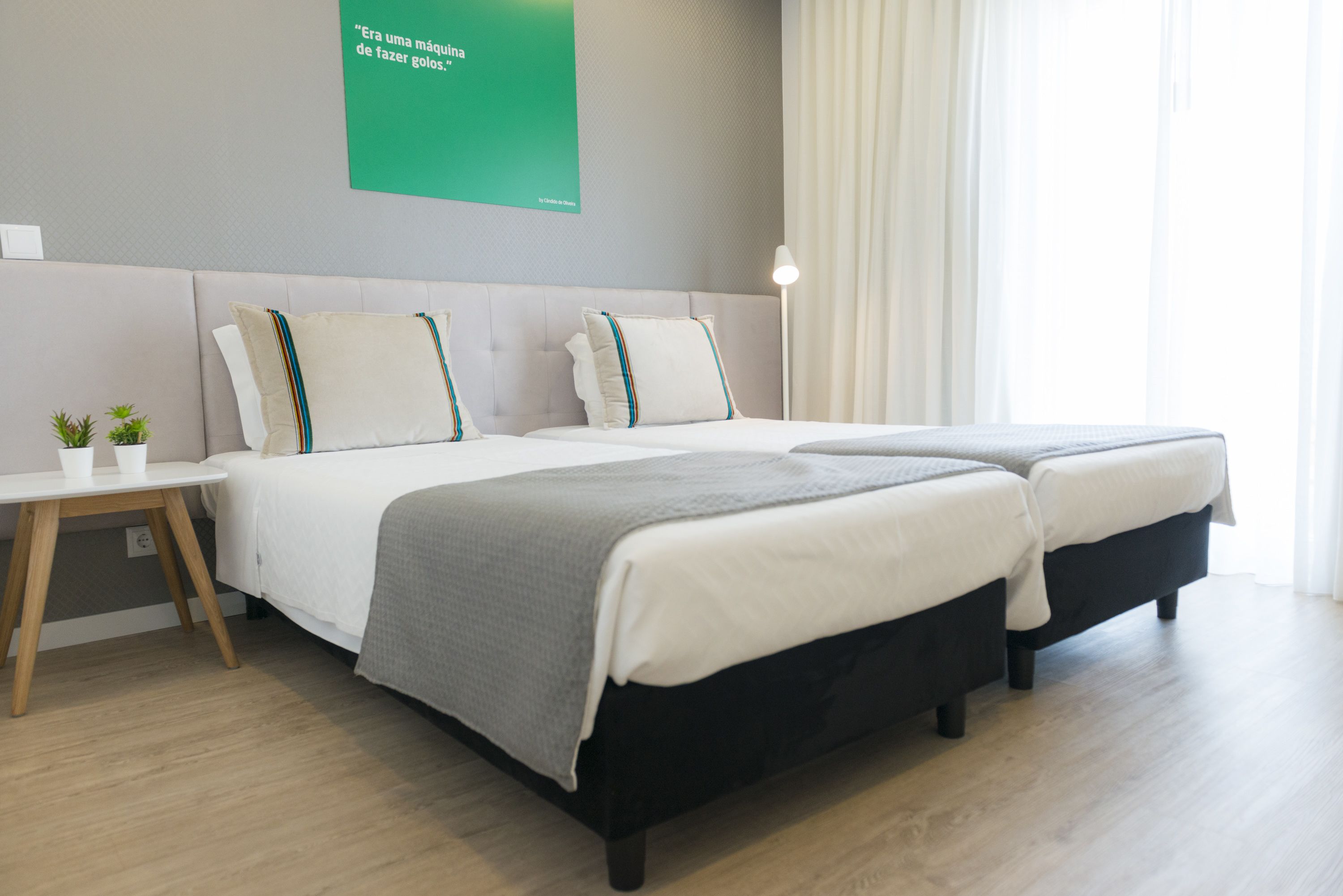 A modern hotel room with two beds, a small table with two cups, a green sign on the wall, and a window with curtains.