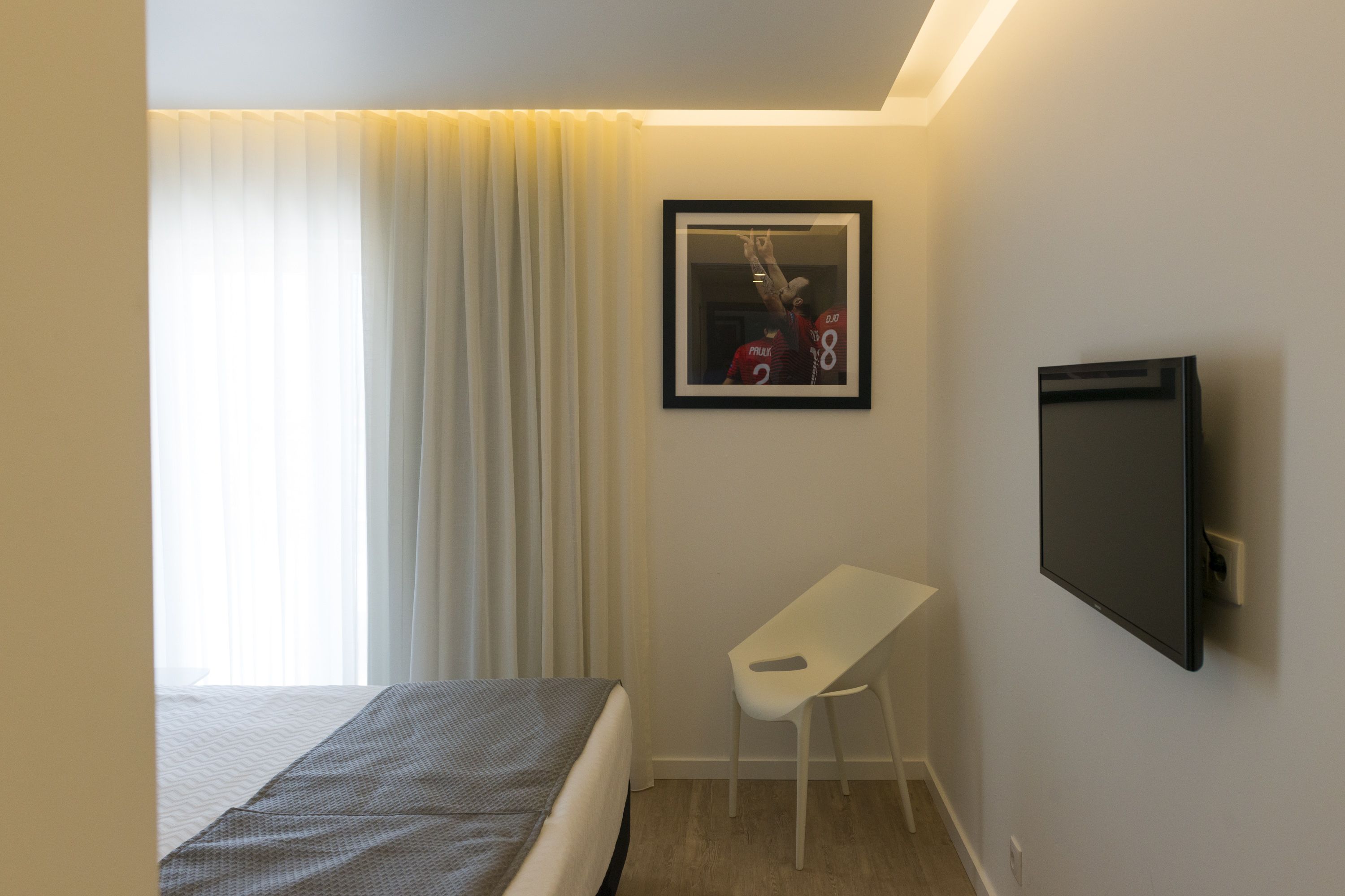 A hotel room with a bed, a white desk, a framed picture on the wall, and a TV mounted on the wall.