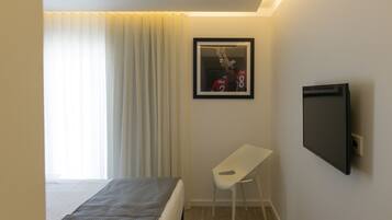 A hotel room with a bed, a white desk, a framed picture on the wall, and a TV mounted on the wall.