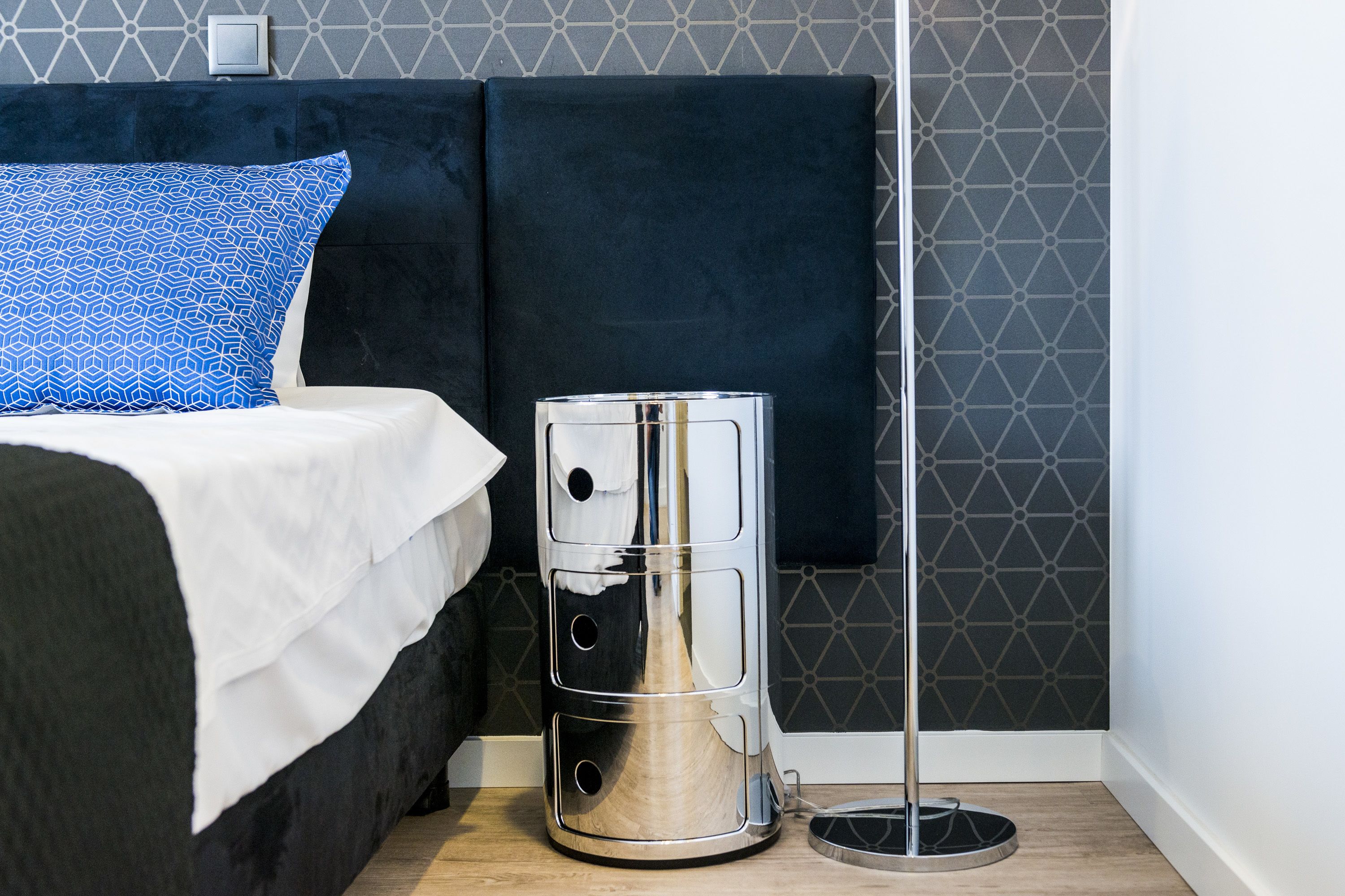 A bed with a white bedspread and a blue pillow, a black headboard, and a metallic trash can with a circular base.