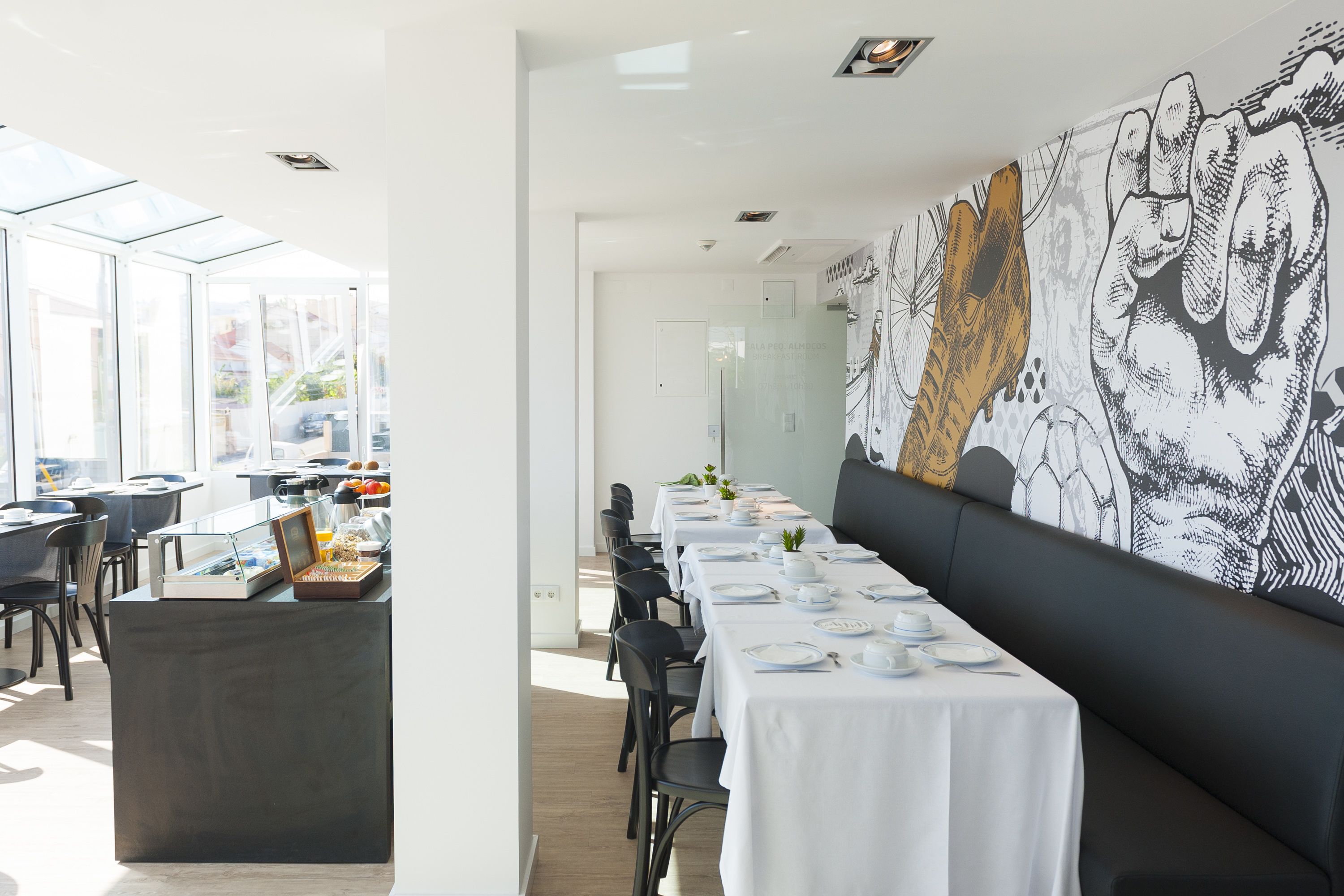 A modern dining area with long tables set for a meal, a black cushioned bench, and a large wall mural.