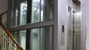 Un ascenseur moderne doté de portes en verre et d’un écran numérique, situé à côté d’un escalier avec une rampe en bois.