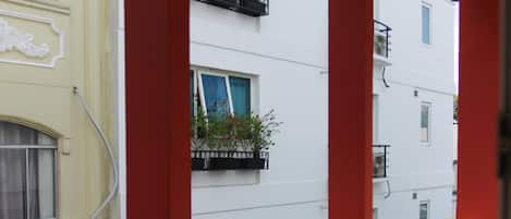 A balcony with a red railing, potted plants, and a view of adjacent buildings.