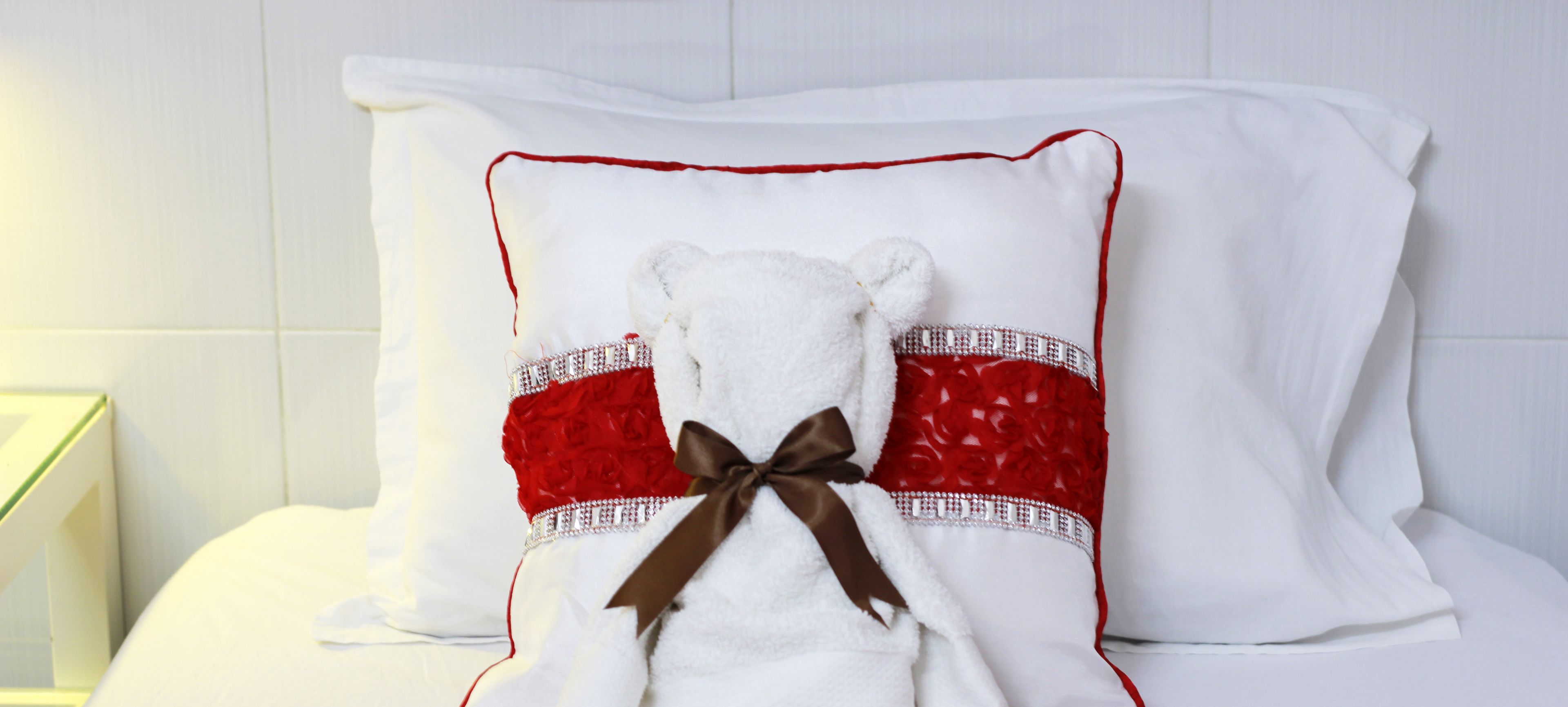 A bed with a white pillow featuring a red and white design, a white bear-shaped pillow, and a brown ribbon.