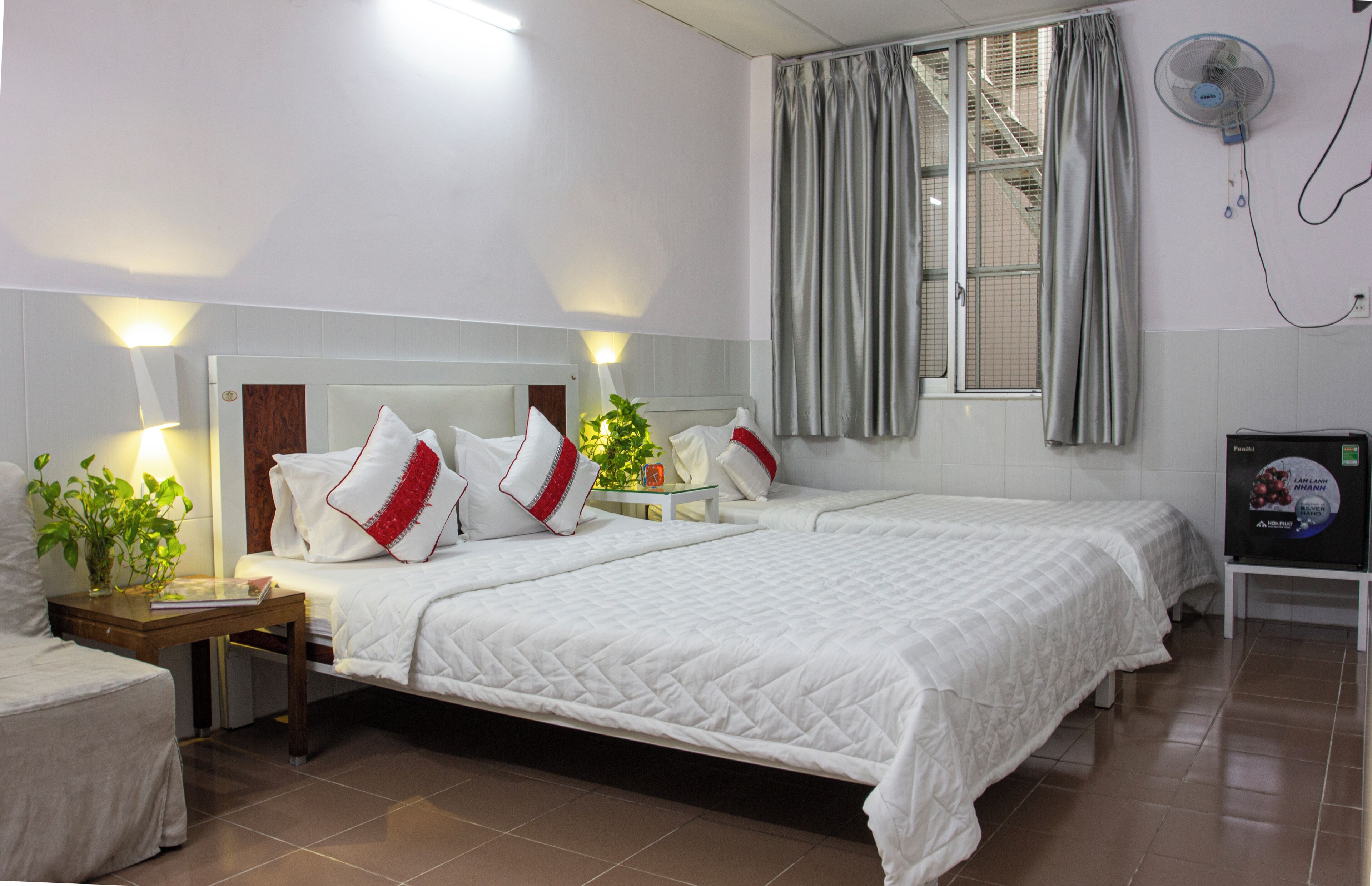 Habitación de hotel con dos camas, una mesita de noche con lámpara, una planta en maceta y un ventilador de pared.