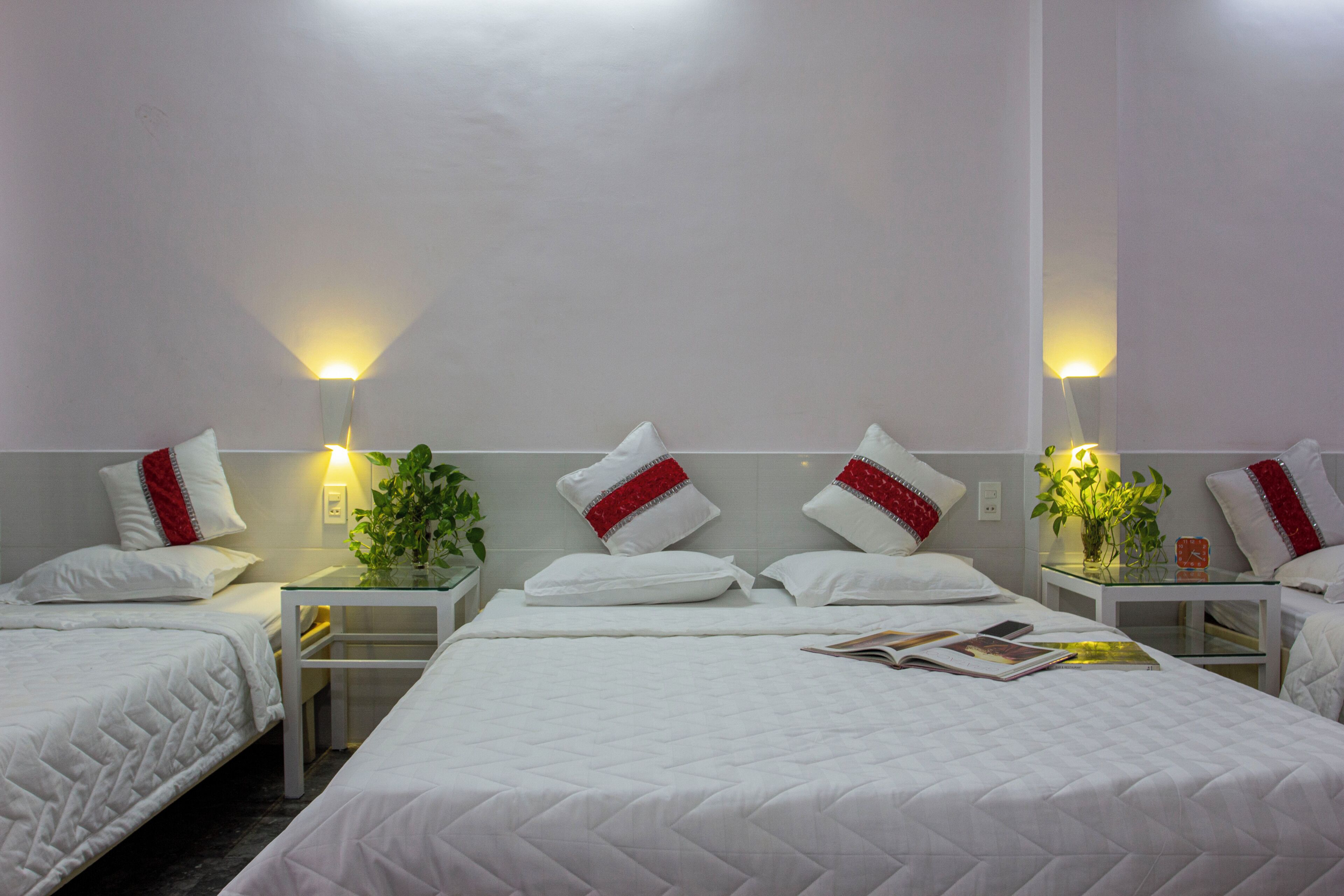A hotel room with two beds, white bedding, red and silver decorative pillows, bedside tables with lamps, and a small plant on one table.