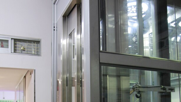 Un ascenseur moderne doté de portes en verre et d’une structure métallique, qui donne sur un hall d’entrée avec une entrée bien visible et un tapis vert à la base.