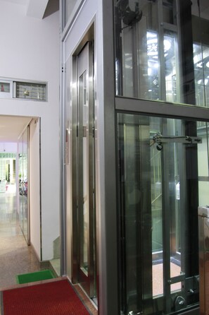 A modern elevator with glass doors and a metal frame, leading to a lobby area with a visible entrance and a green mat at the base. - Quang Saigon Hotel (Ho Chi Minh City)