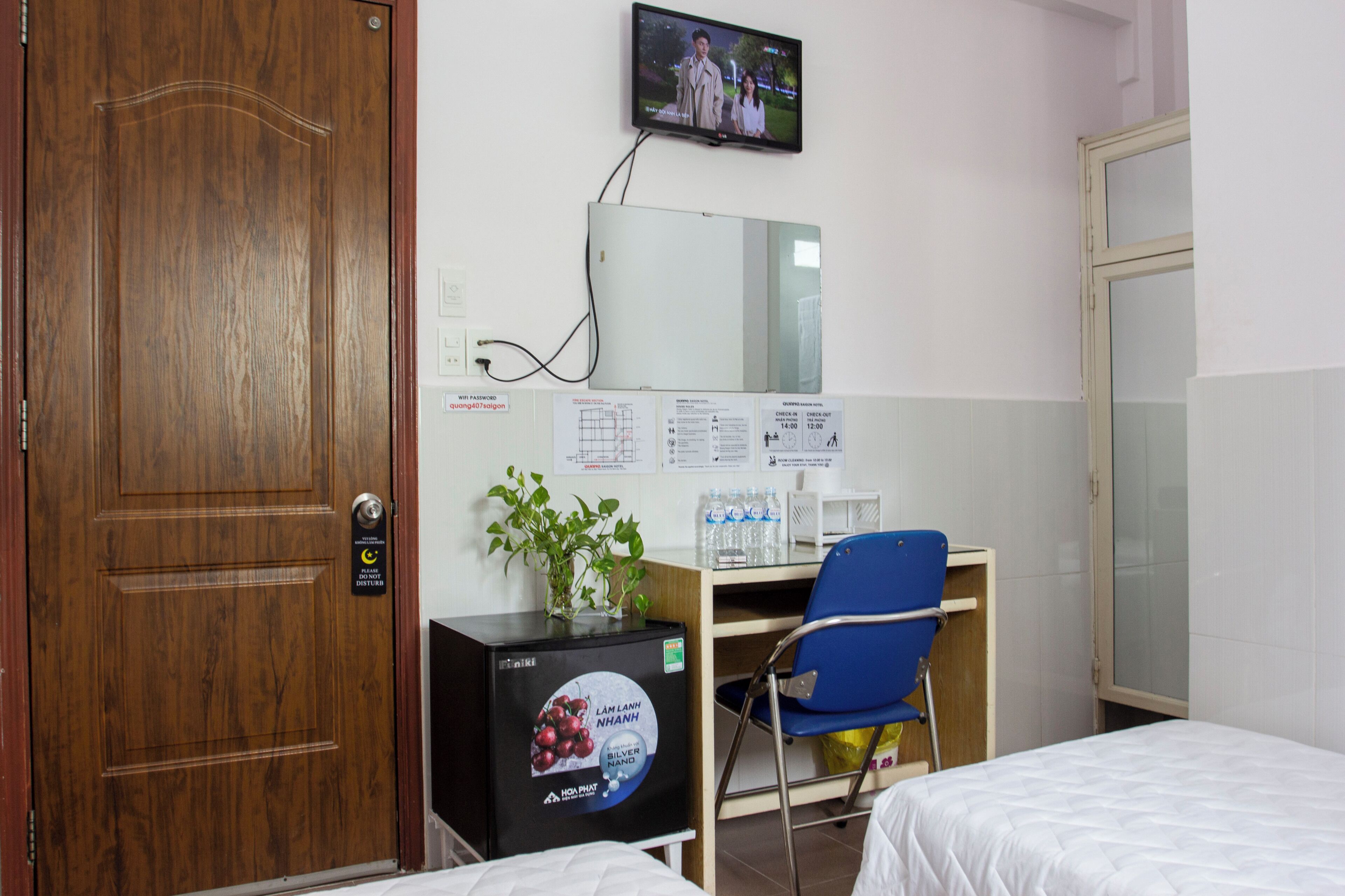 A hotel room with a wooden door, a desk with a computer, a television mounted on the wall, and a bed.