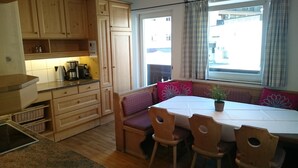 A compact kitchen with wooden cabinets, a dining table with chairs, and a window with curtains.