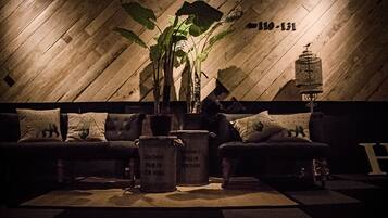 Een gedempt verlichte kamer met een houten wand, een bank met kussens, een potplant en twee metalen bakken met de tekst 'LONDON PARIS NEW YORK'.