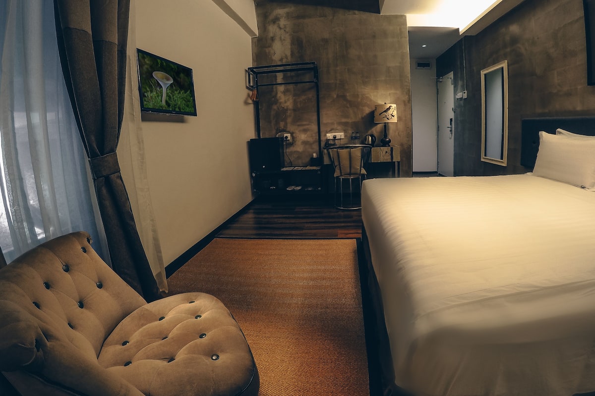 A hotel room with a large bed, a tufted armchair, a desk with a chair, and a television on the wall.